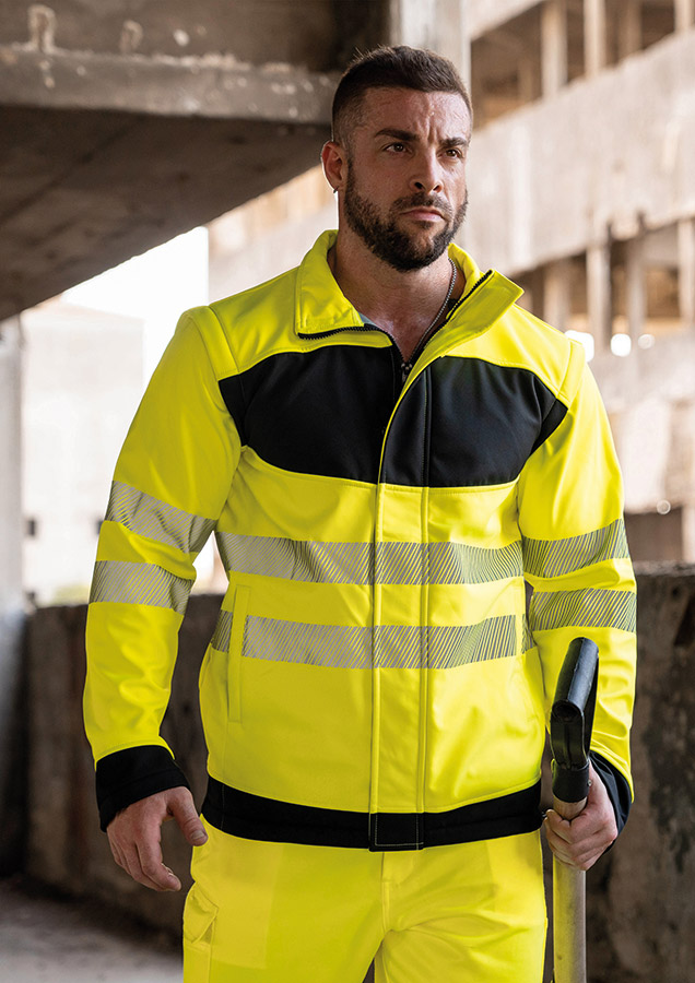 Erwachsener trägt eine gelbe Korntex-Softshell-Jacke mit schwarzer Brustpartie in einem industriellen Umfeld