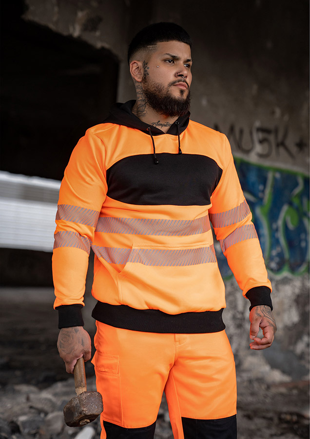 Erwachsener trägt einen orangefarbenen Korntex-Hoodie mit schwarzer Brustpartie in einem industriellen Umfeld