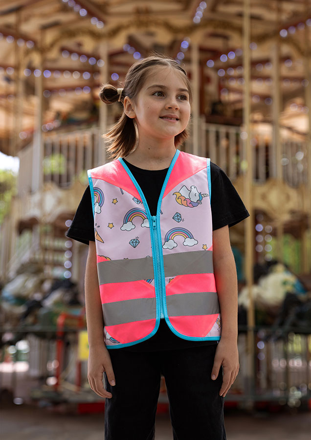 Kind in einer neonpinken Korntex-Warnweste mit Einhorn-Muster vor einem Kinderkarusell