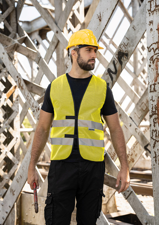 Erwachsener Bauarbeiter steht in gelber Korntex Mesh-Warnweste und gelbem Schutzhelm auf einer Baustelle und hält Werkzeug