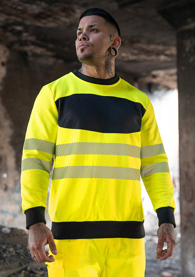 Erwachsener trägt ein gelbes Korntex-Sweatshirt mit schwarzer Brustpartie in einem industriellen Umfeld