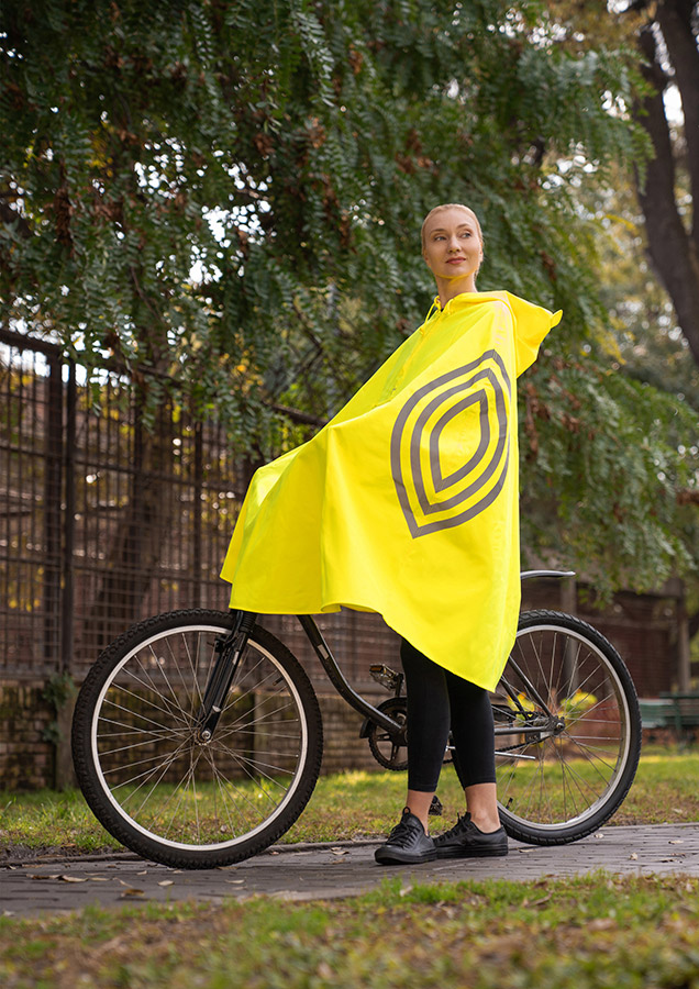 Frau trägt einen gelbfarbenen Regenponcho mit Kapuze, während sie neben einem Fahrrad auf einem Radweg steht