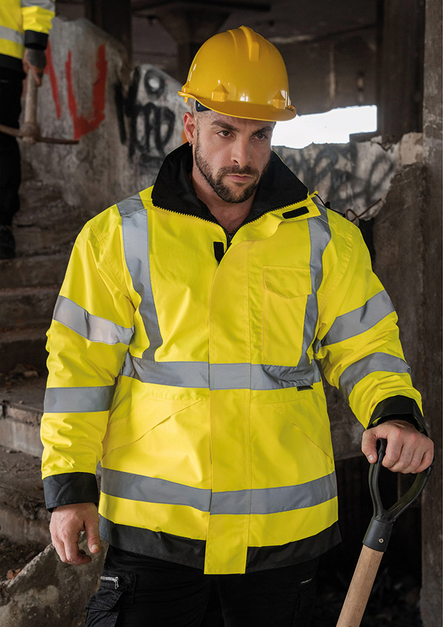 Erwachsener Bauarbeiter in gelber Korntex Hochsichtbarkeits-Regenjacke und gelbem Schutzhelm trägt eine Schaufel auf einer Baustelle.