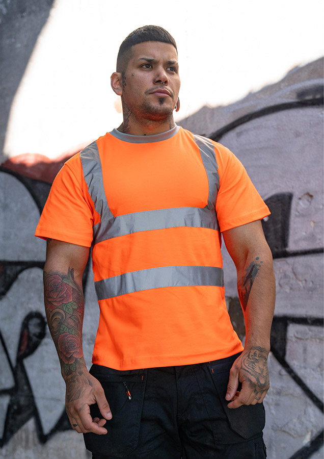 Erwachsener junger Mann in signal-orangenem Korntex T-Shirt mit reflektierenden Streifen steht vor einer Graffiti-Wand
