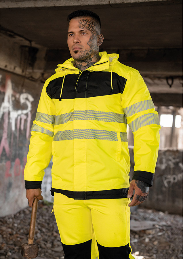 Erwachsener in signalgelbem Korntex-Parka mit schwarzer Brustpartie und Reflexstreifen in einem industriellen Umfeld