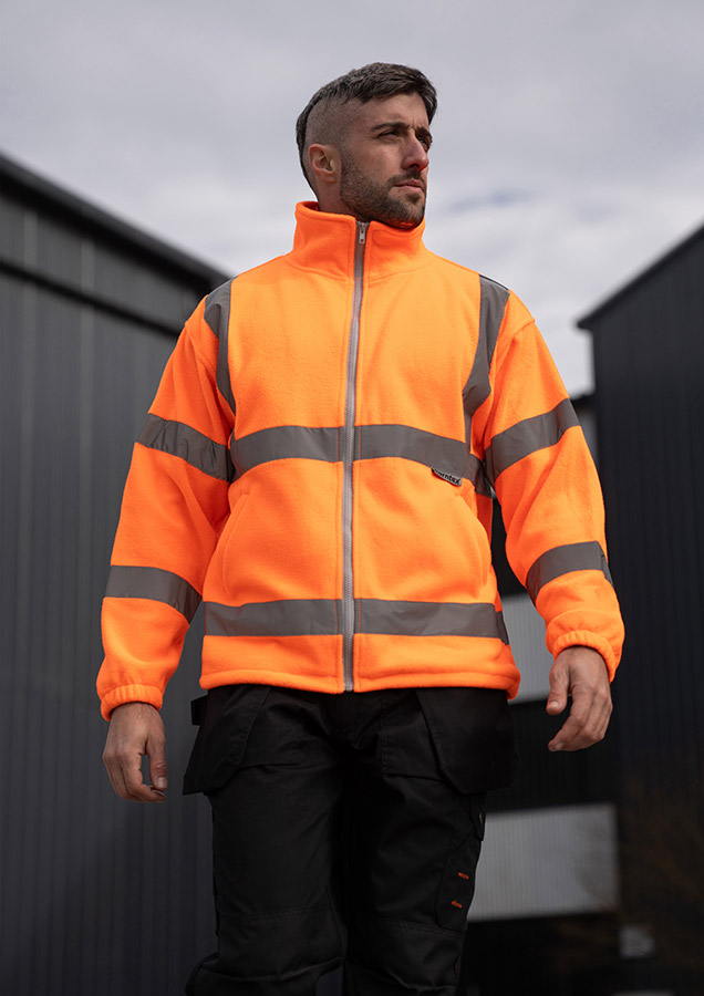 Erwachsener Mann läuft in signal-oranger Korntex Warnschutz-Fleecejacke mit Reflexstreifen vor einem Industriegebäude entlang.