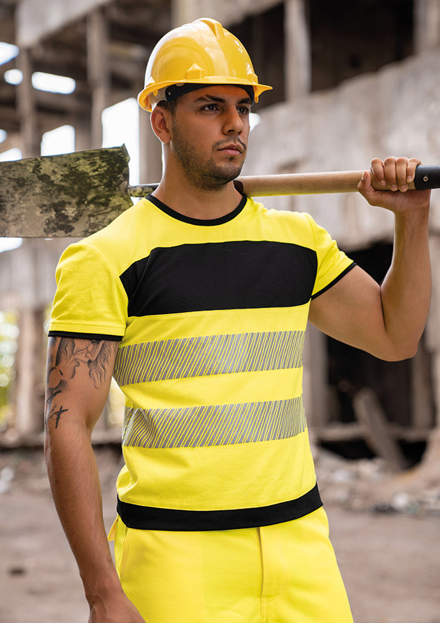 Erwachsener in signalgelbem Korntex-T-Shirt mit schwarzer Brustpartie und Reflexstreifen in einem industriellen Umfeld