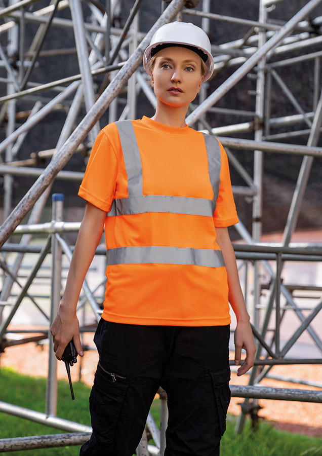 Erwachsene Frau steht in signal-orangenem Korntex Warnschutz-T-Shirt und weißem Schutzhelm vor einem Gerüst auf einer Industriebaustelle