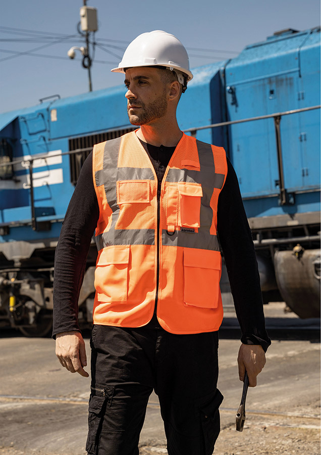 Erwachsener Arbeiter mit orangener Korntex Mesh-Warnweste und Helm hält ein Werkzeug auf einem Bahngelände vor einem Zug
