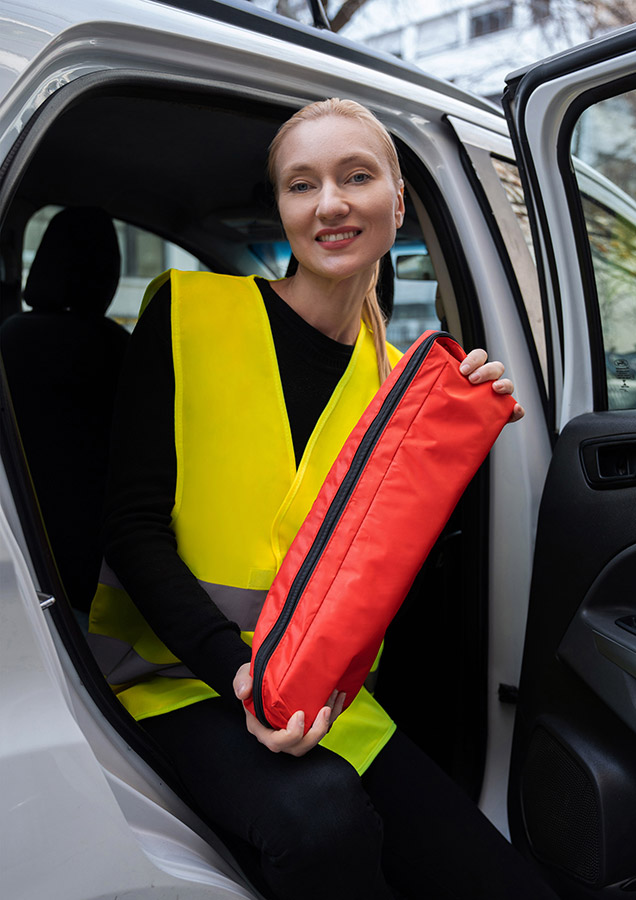 Erwachsene Frau in gelber Korntex Warnweste sitzt im Auto und hält ein rotes Korntex Notfalltaschen-Set