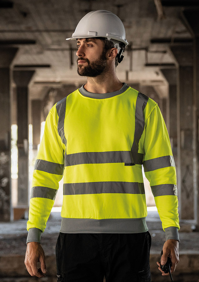 Erwachsener Mann steht in signal-gelbem Korntex Warnschutz-Sweatshirt und weißem Schutzhelm auf einer Baustelle