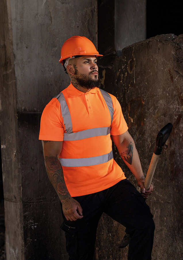 Männlicher junger Arbeiter in einem orangen Korntex Polo-Shirt mit Reflexstreifen hält eine Schaufel in einer baufälligen Umgebung