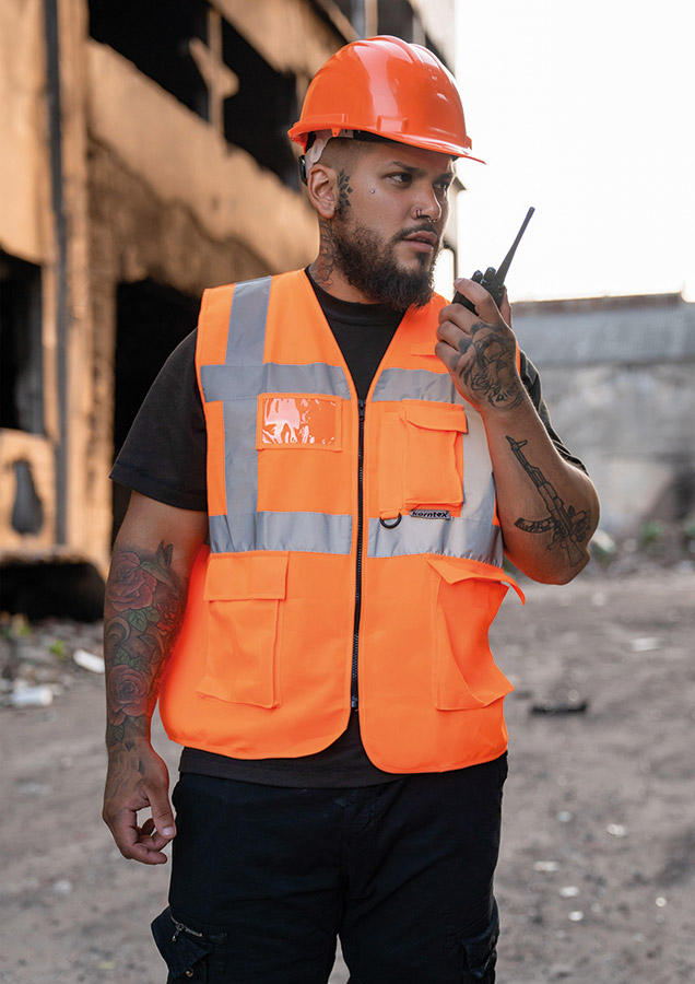 Bauarbeiter in orangener Korntex Multifunktionaler Warnweste und Schutzhelm kommuniziert über Funkgerät auf einer Baustelle