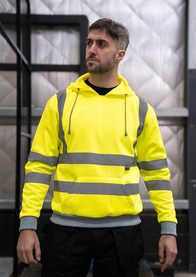 Erwachsener Mann steht in signal-gelbem Korntex Warnschutz-Hoodie mit Reflexstreifen und schwarzer Arbeitshose.