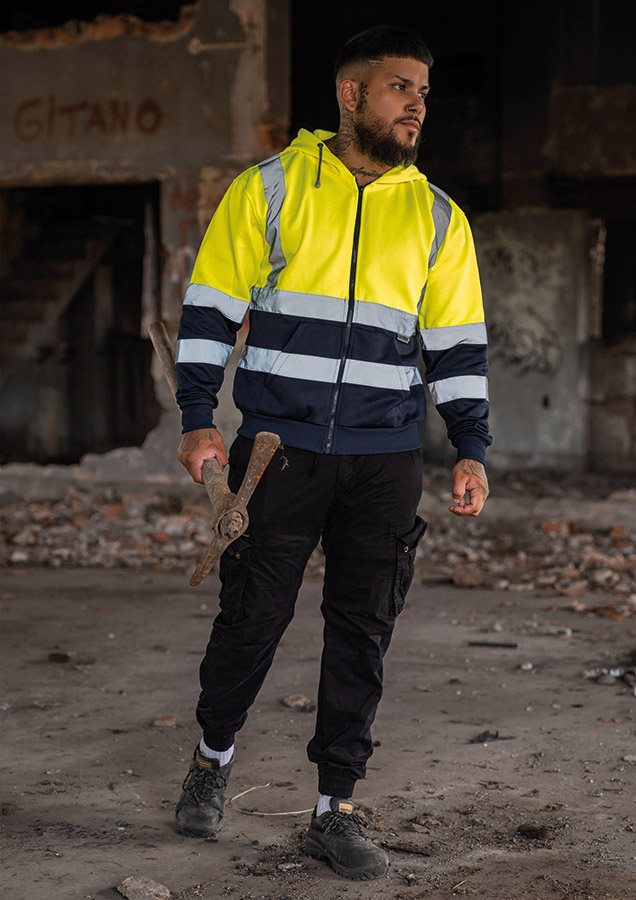 Erwachsener in signalgelbem Korntex-Hoodie mit Zipper und Reflexstreifen in einem industriellen Umfeld
